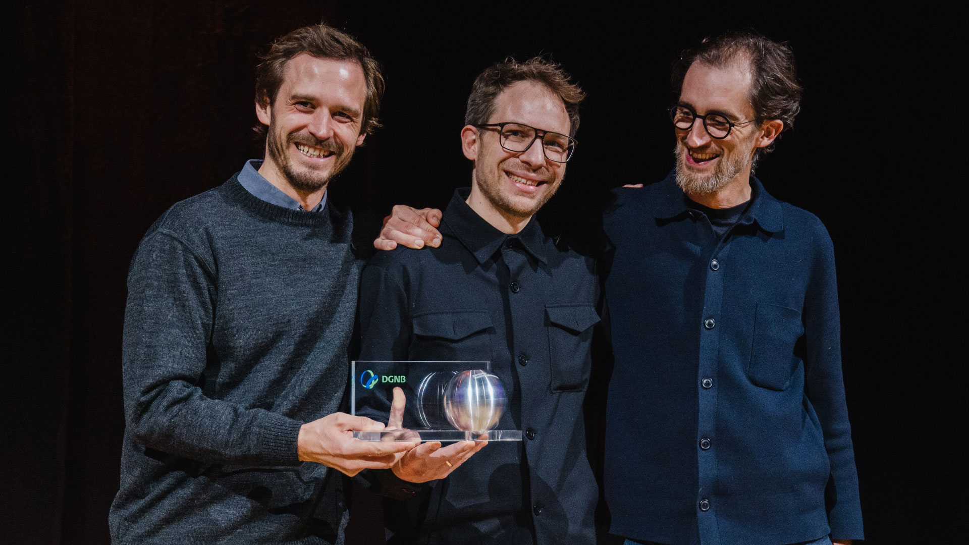 Gruppenfoto von drei Personen nebeneinander vor einem schwarzen Hintergrund. Sie tragen die Preiskugel des Deutschen Nachhaltigkeitspreis gemeinsam in der Hand. Im Bild: Kilian Juraschitz und Florian Kaiser, Atelier Kaiser Shen, Prof. Dr.-Ing. Julian Lienhard, str.ucture