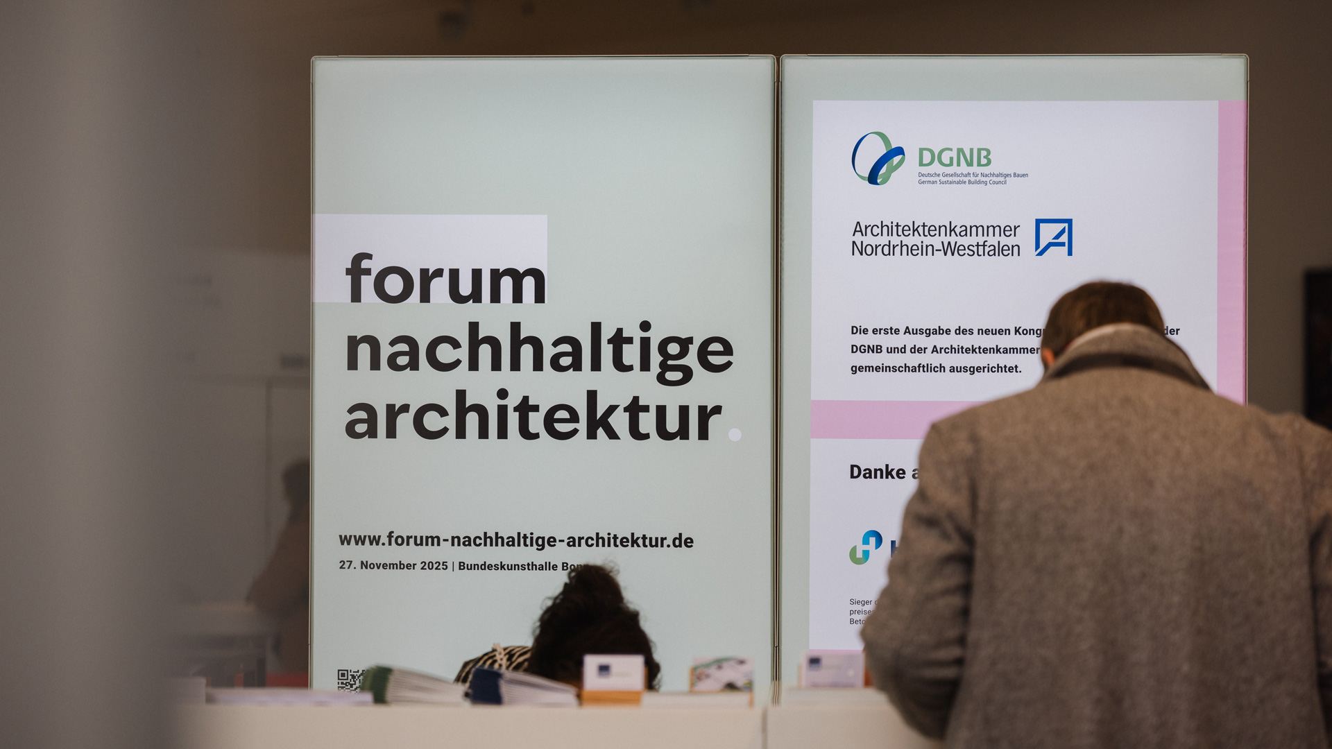 Das Bild zeigt einen Messestand mit dem Logo des Forum nachhaltige Architektur. Davor steht eine Person und betrachtet Informationsmaterialien.